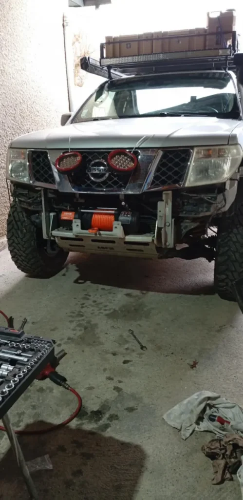 nstallation d'un treuil électrique Vevor 13500 lbs avec corde synthétique orange sur le pare-chocs avant d'un Nissan Patrol blanc en garage, outillage visible au premier plan. Photo issu du site vevor.fr